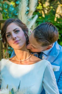 Photographe de mariage 04 - Le marié embrasse le cou de sa femme devant une arche fleurie en Alpes-de-Haute-Provence