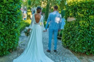 Photographe mariage - Mariés de dos s'avançant dans une allée de jardin, robe de mariée dos nu et costume bleu clair