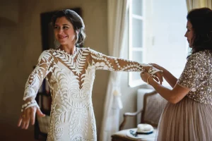 Photographe mariage : Une amie aide la mariée à enfiler sa robe à manches longues dentelle