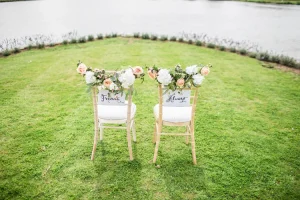 Photographie de mariage : plan sur les chaises des mariés avec panneaux 'Forever' 'Always' et fleurs fraîches, cérémonie en plein air près de l'eau.