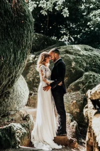 Portrait artistique et romantique des mariés dans un sous-bois de mousse ensoleillé près de Digne-les-Bains. Plan moyen : mariée en robe de dentelle et marié en costume noir, front contre front, capturés par photographe professionnel.
