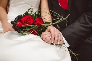 Gros plan sur les mains des mariés avec alliances et bouquet de roses rouges sur la robe blanche