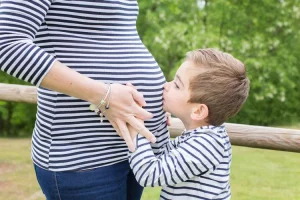 petit frere qui embrasse le ventre de sa maman qui attends un heureux evenement - les deux sont habillés en tenu marine dans un jardin