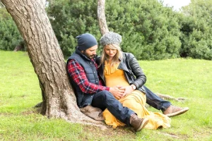 Couple habillé de façon froide en hiver profitant d'un instant pour s'assoir contre un arbre et profiter du ventre rond de sa femme qui attend un heureux évènement
