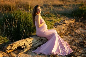 Shooting photo en robe de grossesse type sirene rose pastel posé sur un tronc d'arbre au soleil couchant