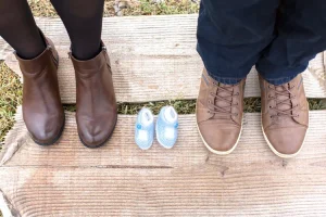 deux paires de pied sur un sol herbeux avec un fond imitations bois et des chaussons de bébé posé au milieu pour annoncer la venu de bébé