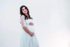 Femme qui sourit largement dans une robe de grossesse - maman se tient le haut et le bas du ventre dans un studio de photographe pro dans les alpes de haute provence