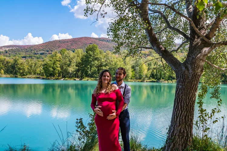 Couple qui attends un heureux evenement maman robe rouge devant un arbre qui baigne dans un petit lac